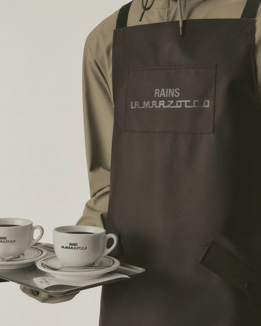 Braune Barista-Schürze von Rains und La Marzocco mit Logo auf der Brusttasche, dazu ein Tablett mit zwei Tassen